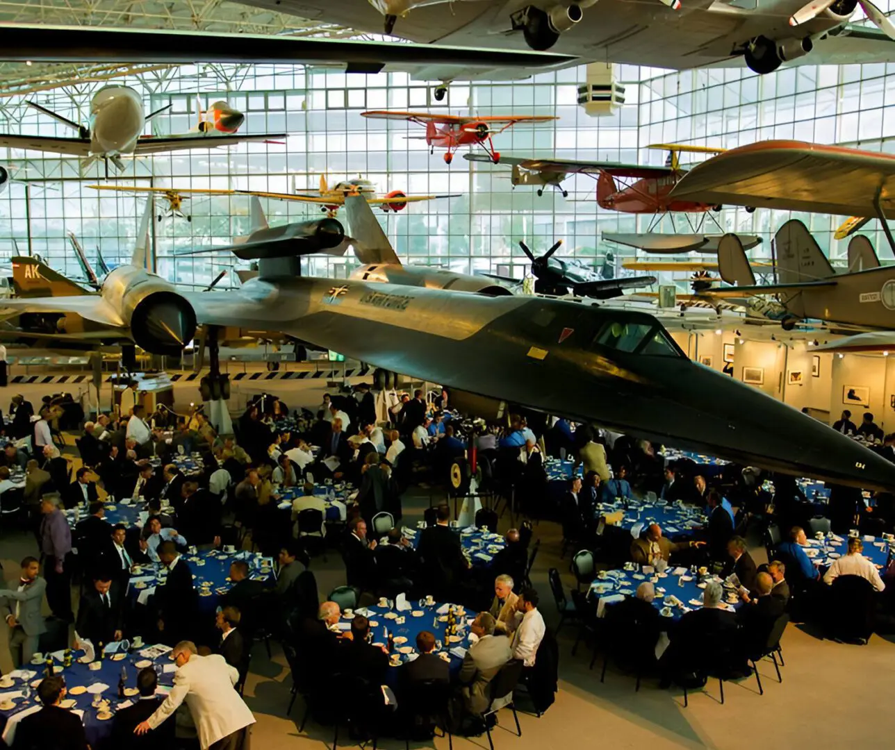 A large group of people sitting at tables in front of airplanes.